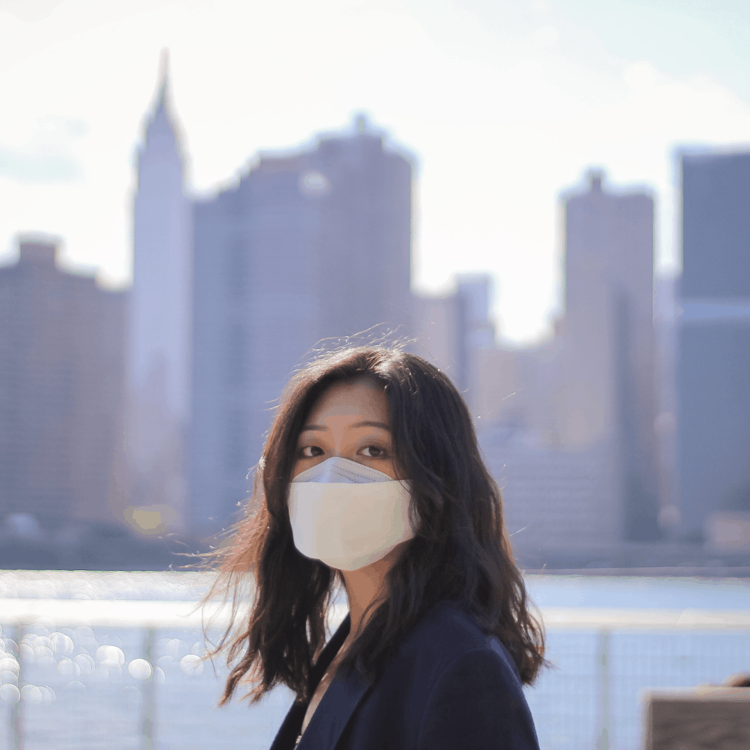 A woman wearing a mask looks at the camera in front of a cityscape.