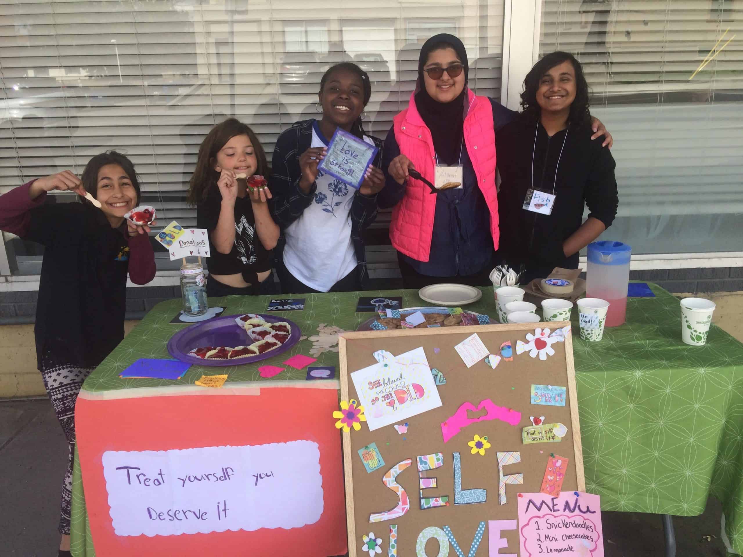 Group of girls standing behind a table with table of refreshments and signs saying "self-love"